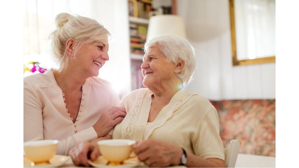 Senior woman & her daughter sitting in living room together, embracing & smiling over tea.
